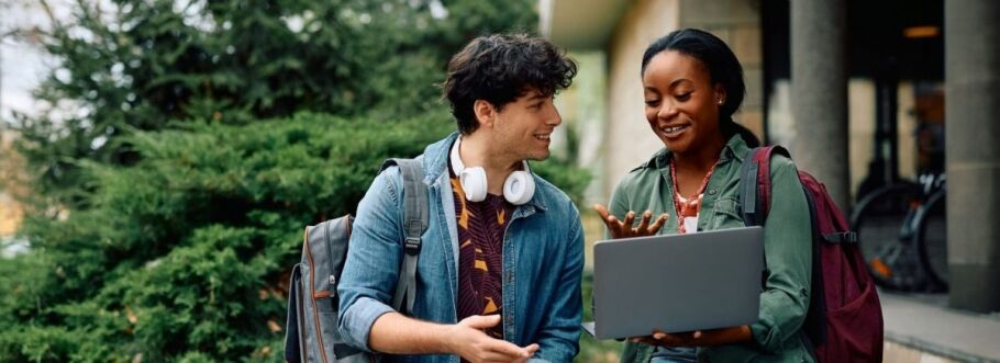 Two college students collaborate on a laptop outdoors near campus.