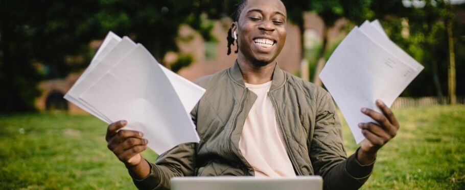 Student smiles, holding college acceptance letters with laptop in foreground.