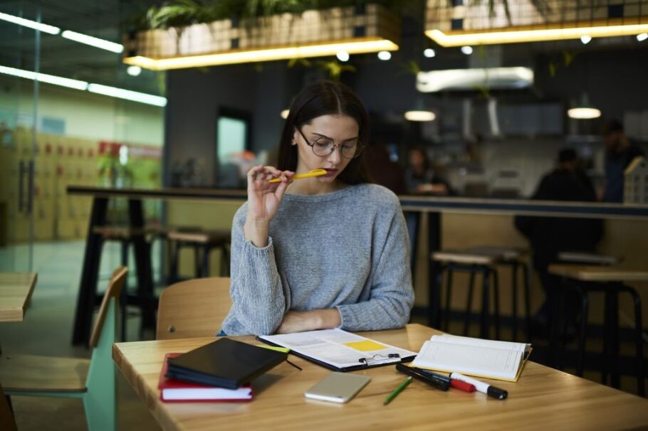 Student considers college application essays, pen in hand, papers on desk.