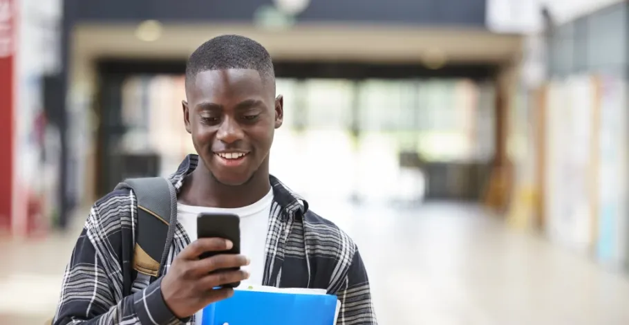 Smiling student checks phone in school hallway, carrying books and backpack.