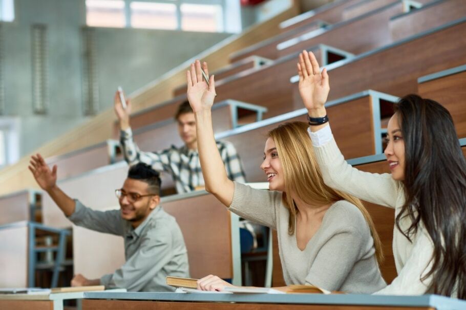 Students in tiered lecture hall raising hands, eager and attentive under bright classroom lighting.