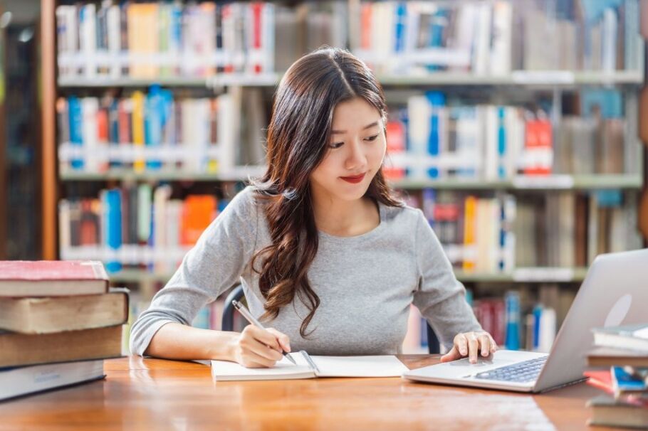 Student researches college options using a laptop and notebook in the library.