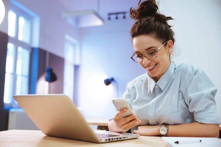 Smiling student checks phone near laptop, possibly about college applications.