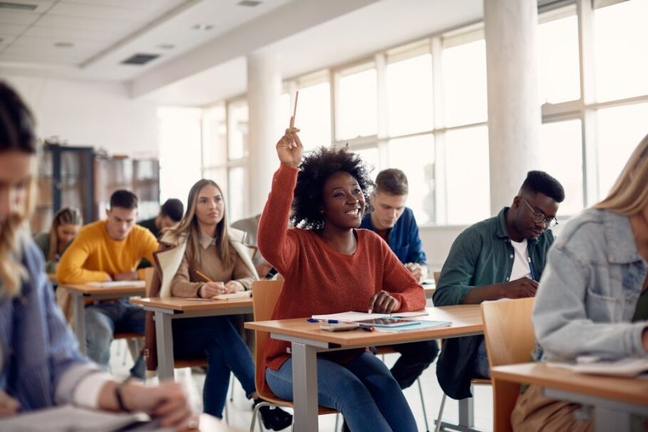 College students in a classroom, one raising hand to ask question.