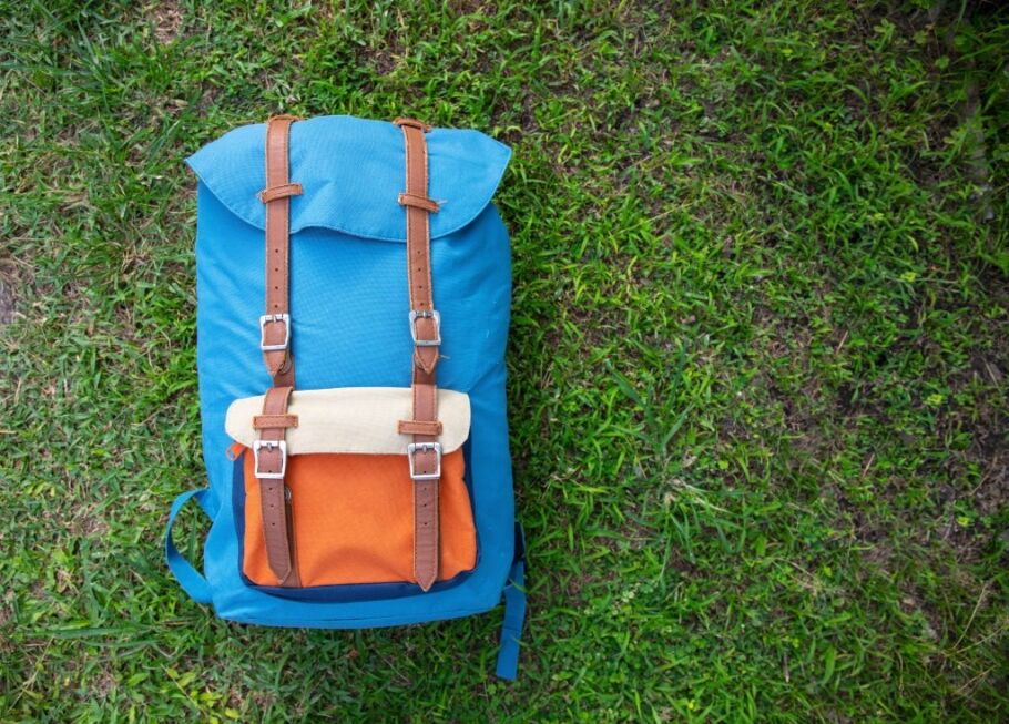 Hipster backpack on green grass. Summer travel prop. Outside trip gear. Blue orange sack on grassland. Forest camp trip accessory. Packing bag for gap year journey. Unisex backpack banner template