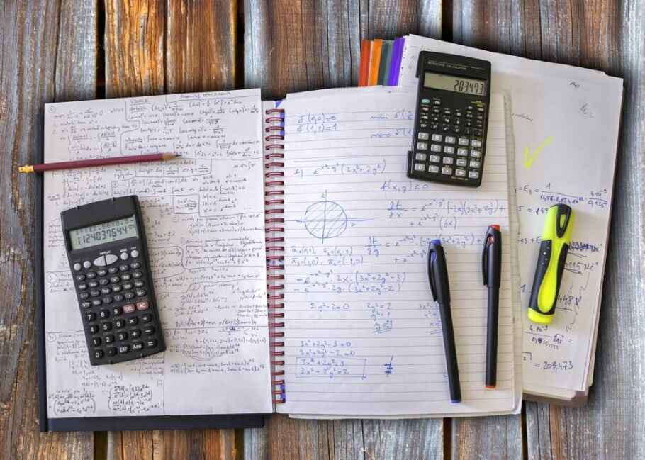 Notebooks, calculators, pens, and papers covered in math equations on a wooden desk.