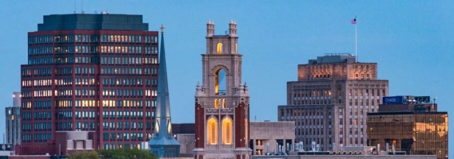 City skyline featuring buildings, a steeple, and a tower against a blue sky.