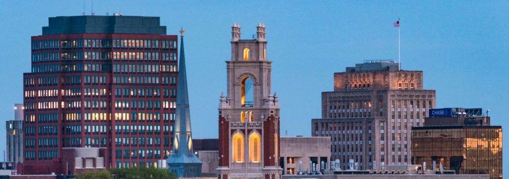 City skyline featuring buildings and a church steeple against a blue sky.
