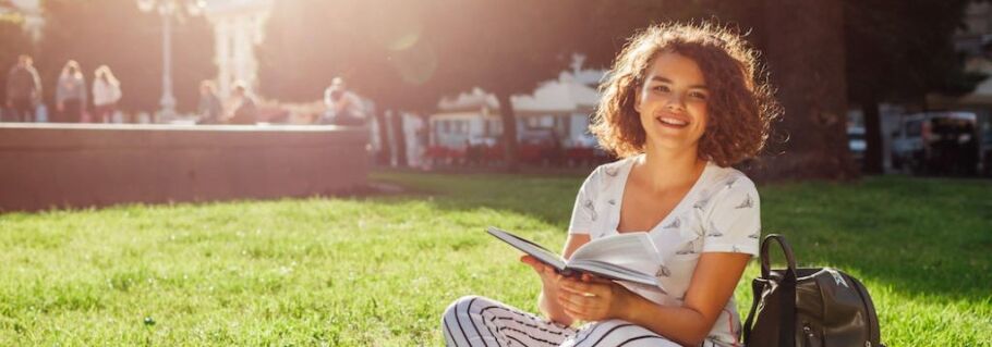 Smiling student reading a book on the grass, with backpack.