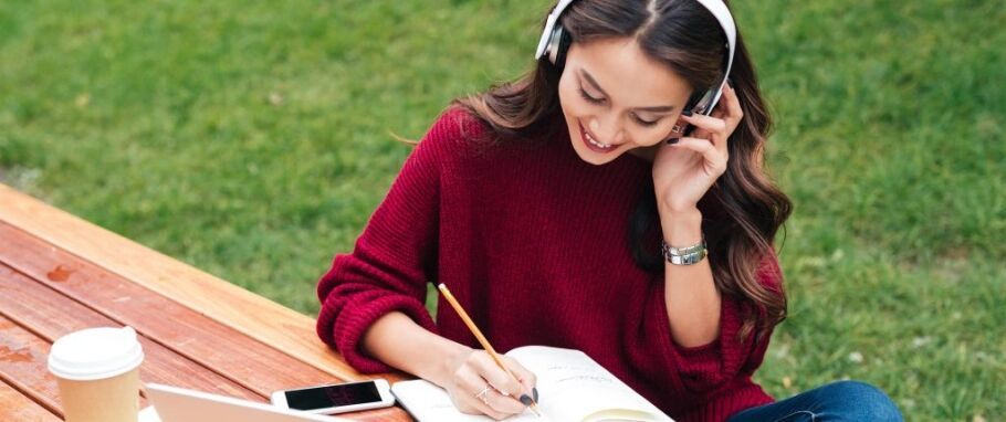 Student with headphones studies outdoors, writing in notebook.