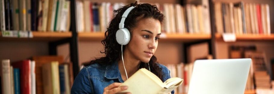 Student with headphones studies with a book and laptop in library.