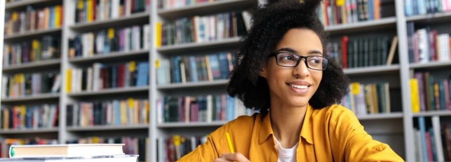 Smiling student with glasses studies in library, thinking about college applications.
