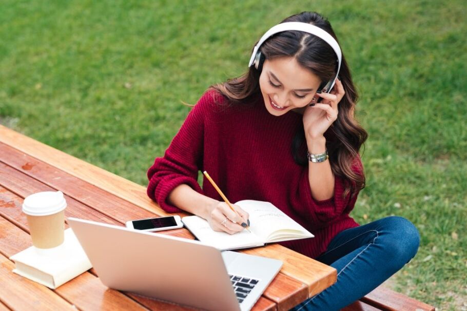 Student on phone, writing in notebook outdoors; college application brainstorming.
