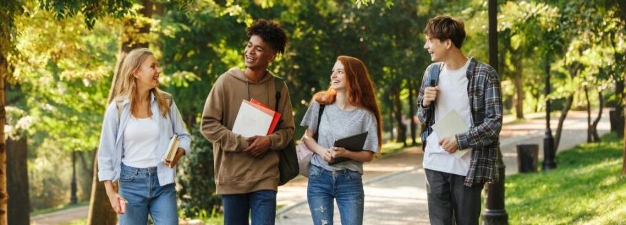 Four smiling students walk on a tree-lined path with books and backpacks.