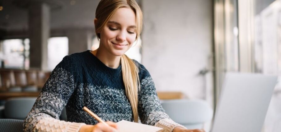 Smiling student studies with laptop and notebook, preparing for college applications.