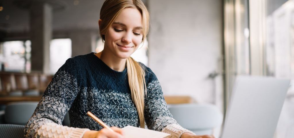 Smiling student studies with laptop and notebook, preparing for college applications.