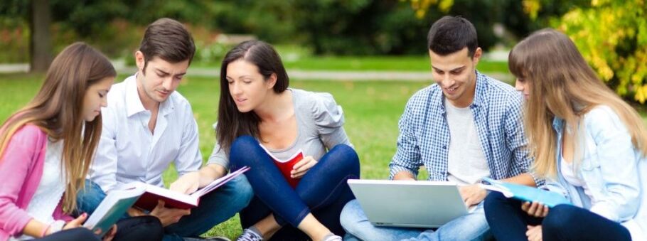 Students seated on grass with laptops and textbooks engaged in focused group study.