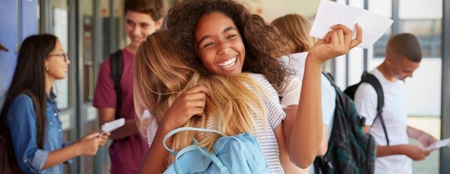 Excited students hug, holding acceptance letters in a school hallway.