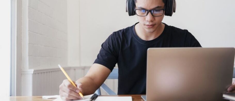 Student wearing headphones studies with laptop and notebook.