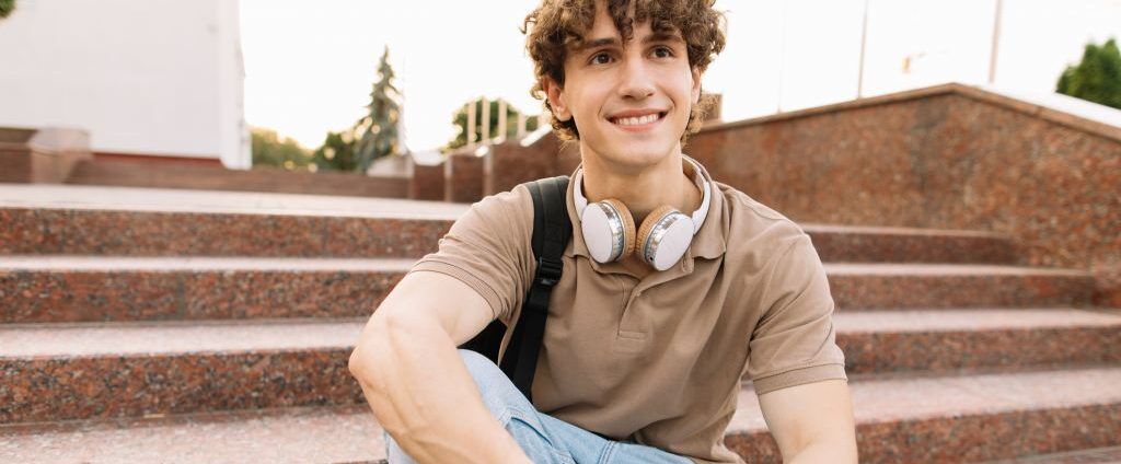 Smiling student with headphones sits on steps, ready for college success.