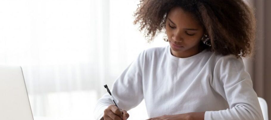 Student writing college application essay with laptop nearby.