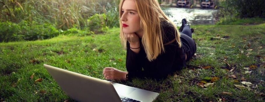 Student with laptop lying on grass, contemplating college application essays.