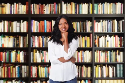 Smiling woman in front of a bookshelf, representing academic knowledge.