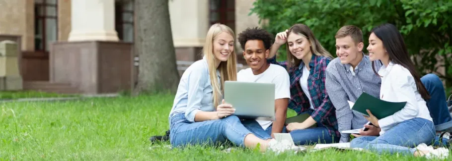 Smiling students collaborate on a laptop outdoors on a college campus lawn.