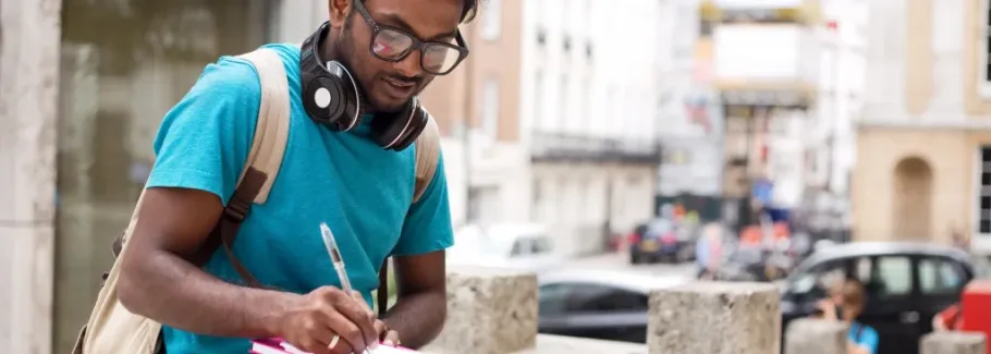 Student with backpack and headphones takes notes outdoors.