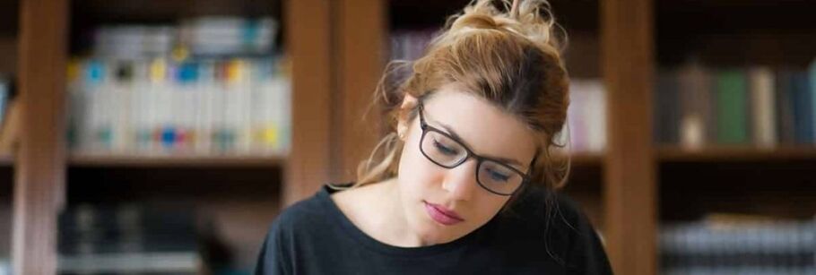 Student wearing glasses studies with books in background.