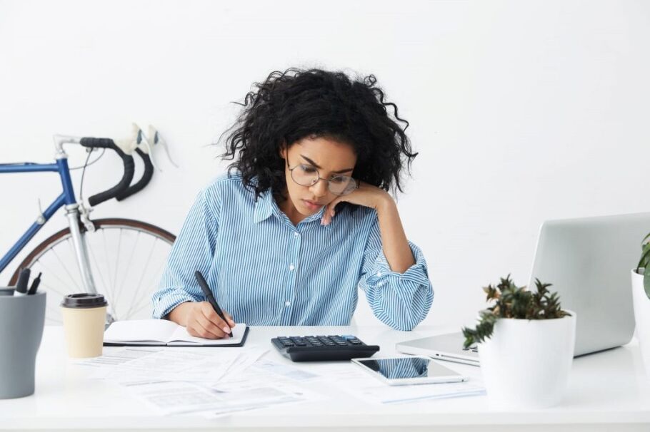 Candid shot of confident hardworking young businesswoman with curly hair sitting at her desk in white office