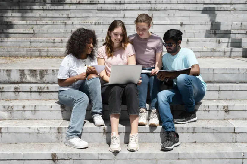 Diverse students collaborate on college applications, sitting on stone steps with a laptop.