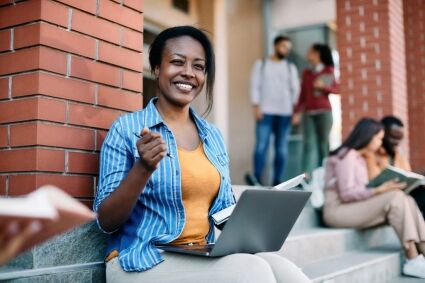 Smiling student with laptop and notebook on campus steps.