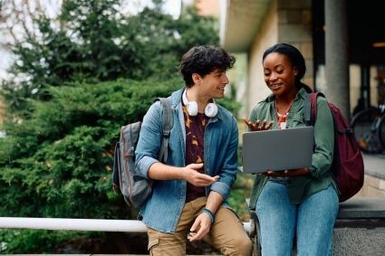 Two students collaborate on a laptop outdoors on a college campus.