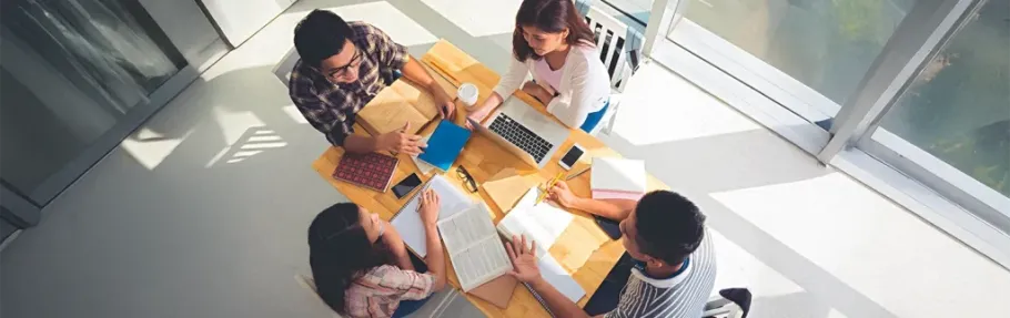 Students collaborate around a table with books and a laptop.