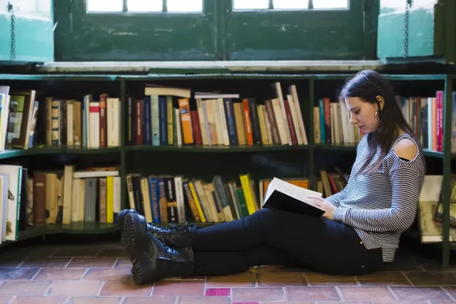 Student reading a book on the floor in front of a bookshelf.