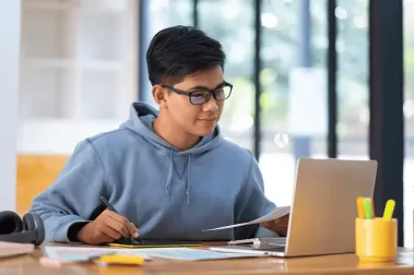 High school student focused on a laptop while holding a pen and paper, ready for college admissions advising.