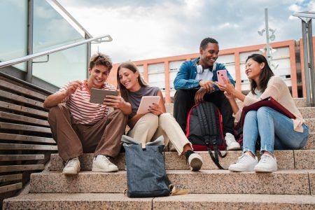 Smiling students using tablets and phones sit on steps outside a college building.